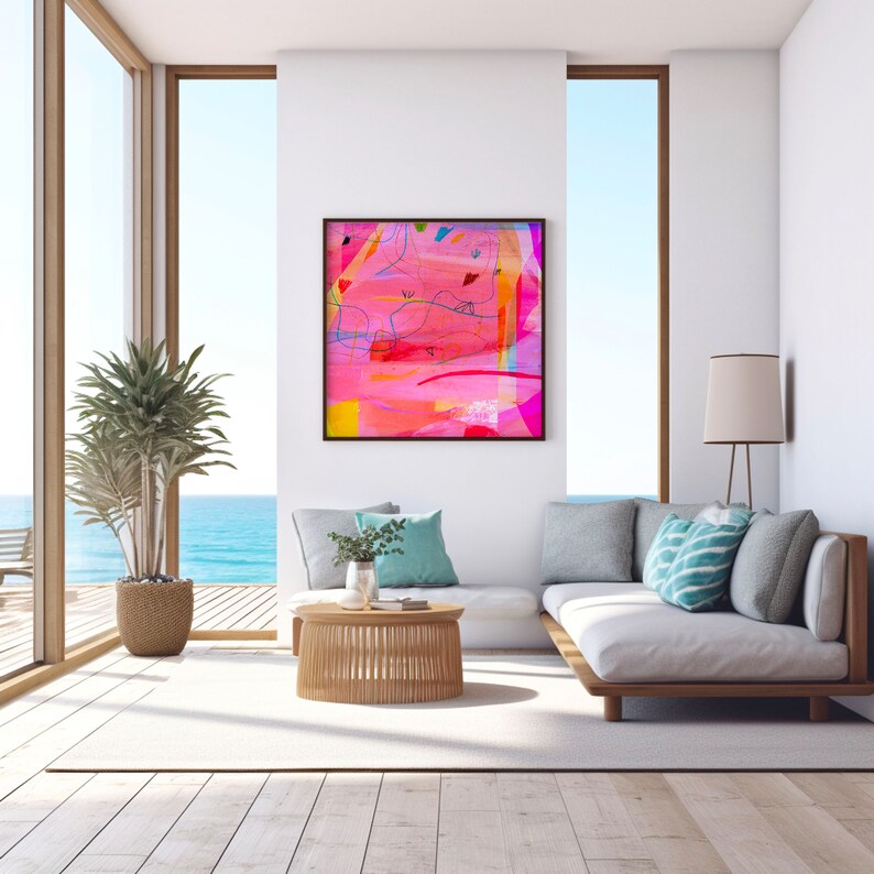 May include: A white couch with blue and white pillows sits in a living room with a large window overlooking the ocean. A framed abstract painting with pink, yellow, and blue colors hangs on the wall above the couch. A wooden coffee table sits in front of the couch.
