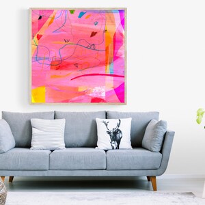 May include: A framed abstract painting with pink, yellow, blue, and red colors. The painting is in a wooden frame and is hanging on a white wall above a gray sofa.