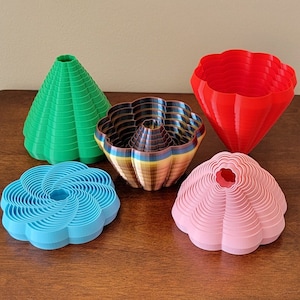 Puede incluir: Cinco cuencos con forma de flor impresos en 3D en diferentes colores. Los cuencos están hechos de plástico y tienen un diseño en capas. Los colores son verde, marrón, rojo, rosa y azul.