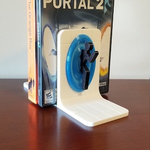 May include: White 3D printed bookend featuring a blue portal and a black figure from the video game Portal 2. The bookend is holding up two video game cases, one for Portal 2 and one for The Orange Box.