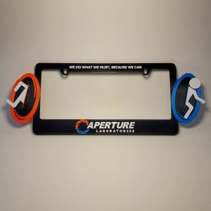 May include: Black license plate frame with a blue and orange portal logo and the text "Aperture Laboratories" and the phrase "We do what we must, because we can".