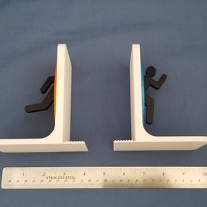 May include: A pair of white bookends shaped like portals from the video game Portal. The portals are orange and blue, and each has a black silhouette of a person running through it.