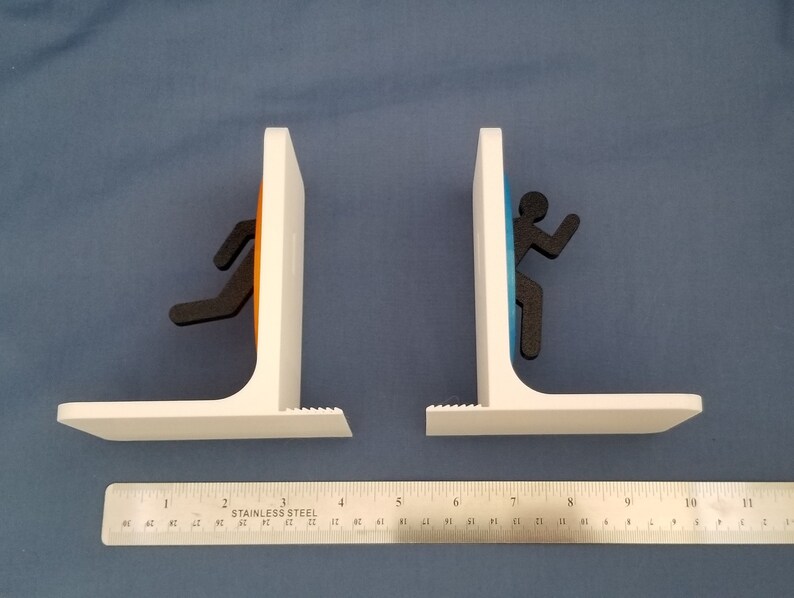 May include: A pair of white bookends shaped like portals from the video game Portal. The portals are orange and blue, and each has a black silhouette of a person running through it.