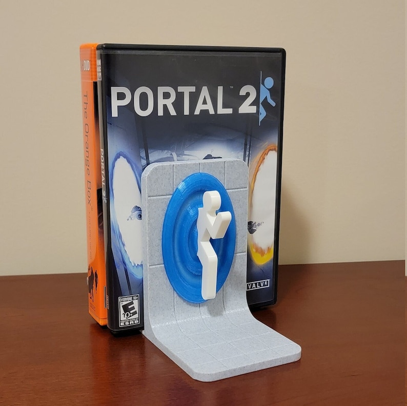 May include: A blue and white 3D printed bookend featuring the Portal 2 logo and a stylized figure. The bookend is designed to hold a video game case. The case is for the game Portal 2, and the text "The Orange Box" is visible on the side of the case.