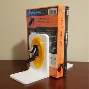 May include: A white 3D printed bookend featuring a portal gun and a yellow portal disc. The bookend is holding up a copy of the video game "The Orange Box" which includes the games Half-Life 2: Episode Two, Portal, and Team Fortress 2. The bookend is also holding up a copy of the video game "Portal 2".