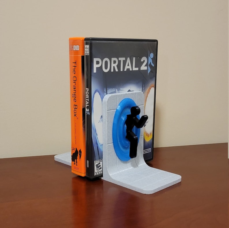 May include: A 3D printed bookend featuring a blue portal and a black figure from the video game Portal 2. The bookend is holding a copy of the Portal 2 video game.