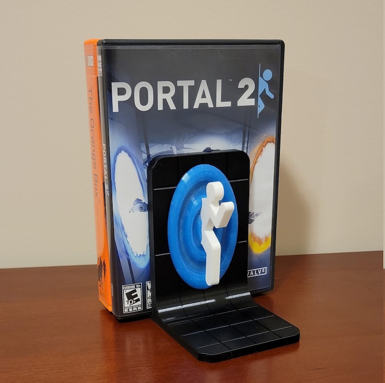 May include: A black bookend featuring a blue portal with a white figure, holding a portal gun, from the video game Portal 2. The bookend is holding up a copy of the Portal 2 video game.