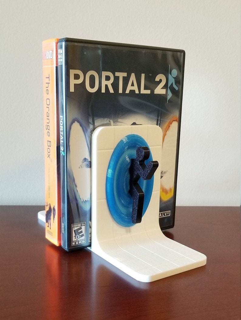 May include: White 3D printed bookend featuring a blue portal and a black figure from the video game Portal 2. The bookend is holding up two video game cases, one for Portal 2 and one for The Orange Box.