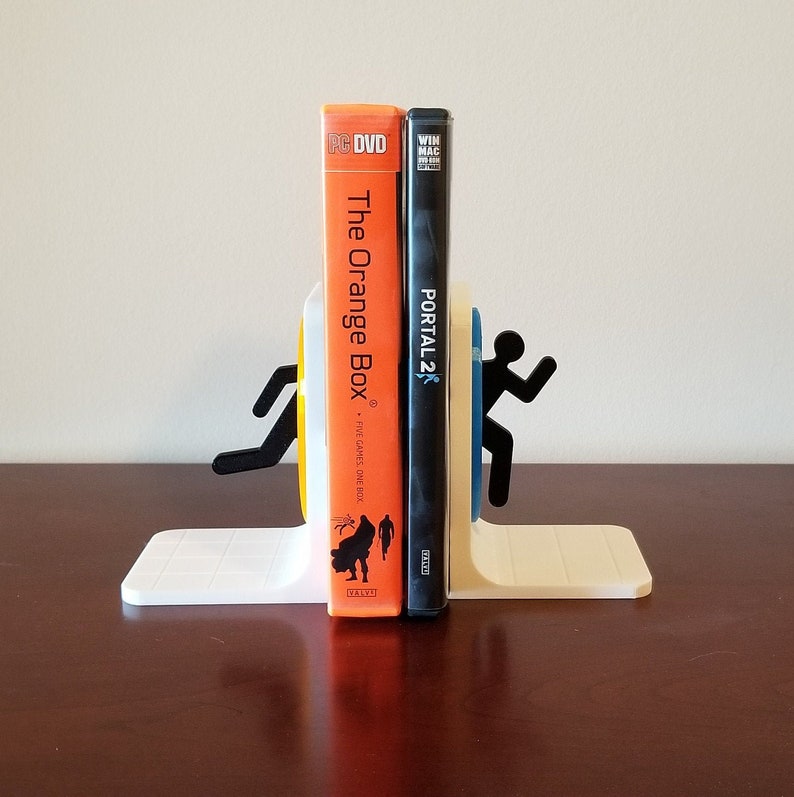 May include: White 3D printed bookends shaped like the Portal 2 characters. The bookends are holding up a copy of the PC DVD game "The Orange Box" and a copy of the PC DVD game "Portal 2".