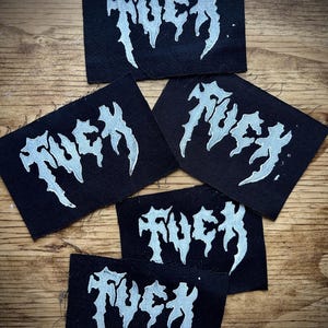 May include: Five black fabric patches with the word "FUCK" in a silver, gothic-style font. The patches are rectangular and arranged on a wooden surface. The text is bold and stylized.