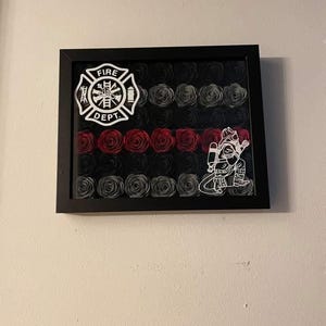 May include: Framed artwork featuring a black background with rows of black, gray, and red paper roses. A white Fire Department emblem and a firefighter graphic are on the frame. The artwork is in a black frame.