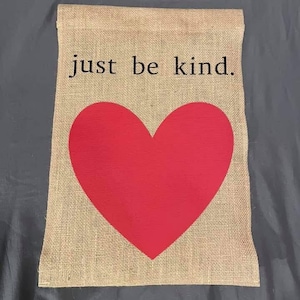Könnte beinhalten: Eine rechteckige Gartenflagge aus Jute mit den schwarzen Worten "just be kind" über einem großen roten Herzen. Die Flagge hat eine natürliche, beige Farbe.