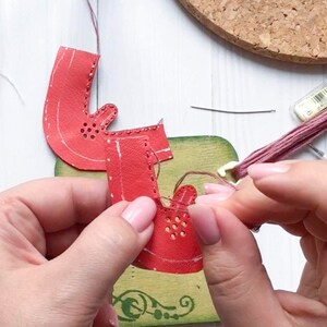 May include: A close-up of a person's hands stitching two red leather pieces together with a brown thread. The leather pieces have a pattern of small holes and are being sewn on a green surface.