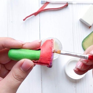 May include: A person is using a paintbrush to apply glue to a red leather shoe. The shoe is being held in place by a green tool.
