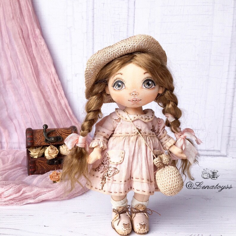 May include: A handmade doll with brown hair in braids, wearing a pink dress, a straw hat, and a crocheted bag. The doll is standing on a white surface with a wooden box and flowers in the background.
