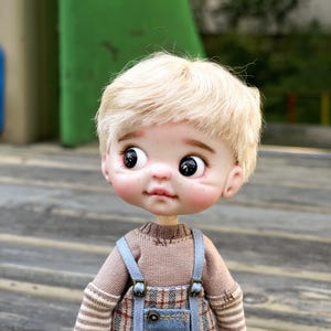 Puede incluir: Una muñeca artesanal con tez clara, cabello rubio y ojos grandes y expresivos. La muñeca viste un suéter marrón, un peto azul con estampado de cuadros y mangas a rayas. Una muñeca de colección.