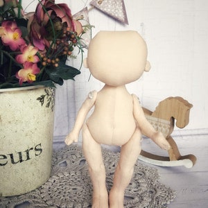 May include: A nude-colored fabric doll with a simple design. The doll has a round head, no facial features, and long limbs. The doll is standing on a crocheted doily.