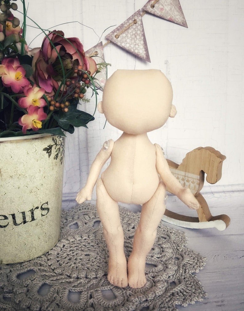 May include: A nude-colored fabric doll with a simple design. The doll has a round head, no facial features, and long limbs. The doll is standing on a crocheted doily.
