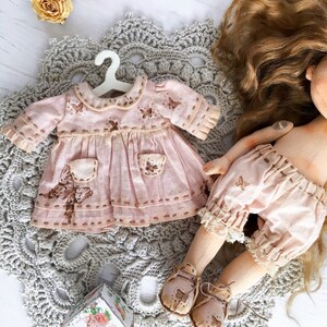 May include: A handmade doll wearing a pink dress with butterfly embroidery and brown shoes. The doll has long brown hair and is sitting on a crocheted doily.
