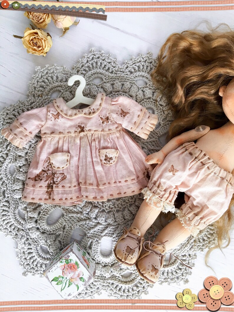 May include: A handmade doll wearing a pink dress with butterfly embroidery and brown shoes. The doll has long brown hair and is sitting on a crocheted doily.
