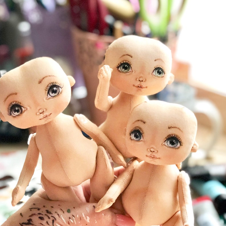 May include: Three handmade cloth dolls with painted faces and large eyes. The dolls are nude and have no clothing. The dolls are posed in a playful manner, with one doll sitting on top of the other two.