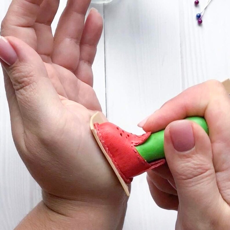 May include: A red leather shoe with a green handle is being pressed onto a piece of beige leather.