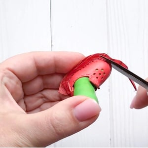 May include: A close-up of a red leather shoe with a green strap being trimmed with scissors. The shoe has a perforated design and is being held by a hand.