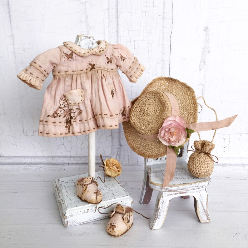 May include: A pink doll dress with butterfly embroidery, a straw hat with a pink flower, and a small crocheted bag on a white wooden chair.