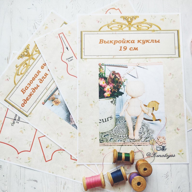 May include: A set of sewing patterns for a 19 cm doll, with the text "Выкройка куклы 19 см" and "Базовая основа одежды для..." on the pages. The patterns are laid out on a white wooden surface with spools of thread in various colors.