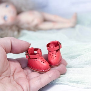 May include: A pair of miniature red leather shoes with buckles. The shoes have a perforated design and are held in a hand.