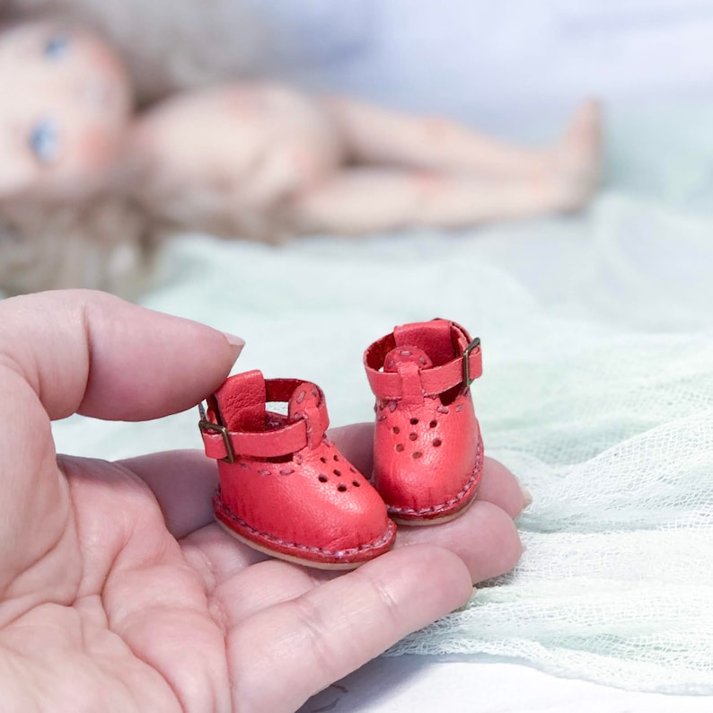 May include: A pair of miniature red leather shoes with buckles. The shoes have a perforated design and are held in a hand.
