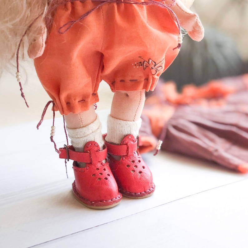 May include: A doll wearing a pair of red leather shoes with a strap and buckle. The doll is wearing a short orange skirt and white socks.