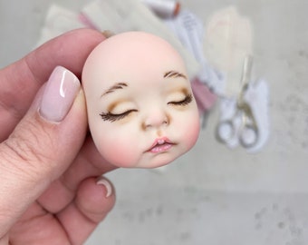 May include: A porcelain doll head with closed eyes, painted brown eyebrows, and a light pink blush on the cheeks.