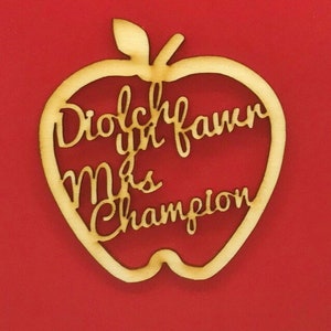 May include: A wooden apple-shaped cutout with the words "Diolch yn fawr Mrs Champion" in a cursive font. The cutout is light brown and set against a red background, suitable for a teacher's gift.