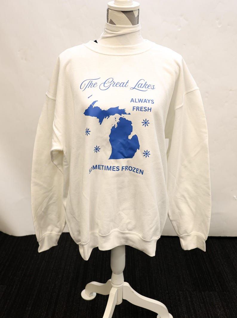 Pu&ograve; includere: Felpa bianca con grafica blu dei Grandi Laghi, compreso lo stato del Michigan. Il testo recita "The Great Lakes, Always Fresh, Sometimes Frozen" con accenti di fiocchi di neve blu. La felpa &egrave; esposta su un manichino bianco.