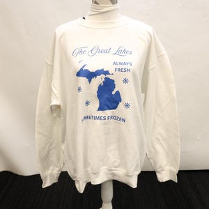 Pu&ograve; includere: Felpa bianca con grafica blu della regione dei Grandi Laghi e la scritta "The Great Lakes ALWAYS FRESH SOMETIMES FROZEN". La felpa &egrave; esposta su un manichino.