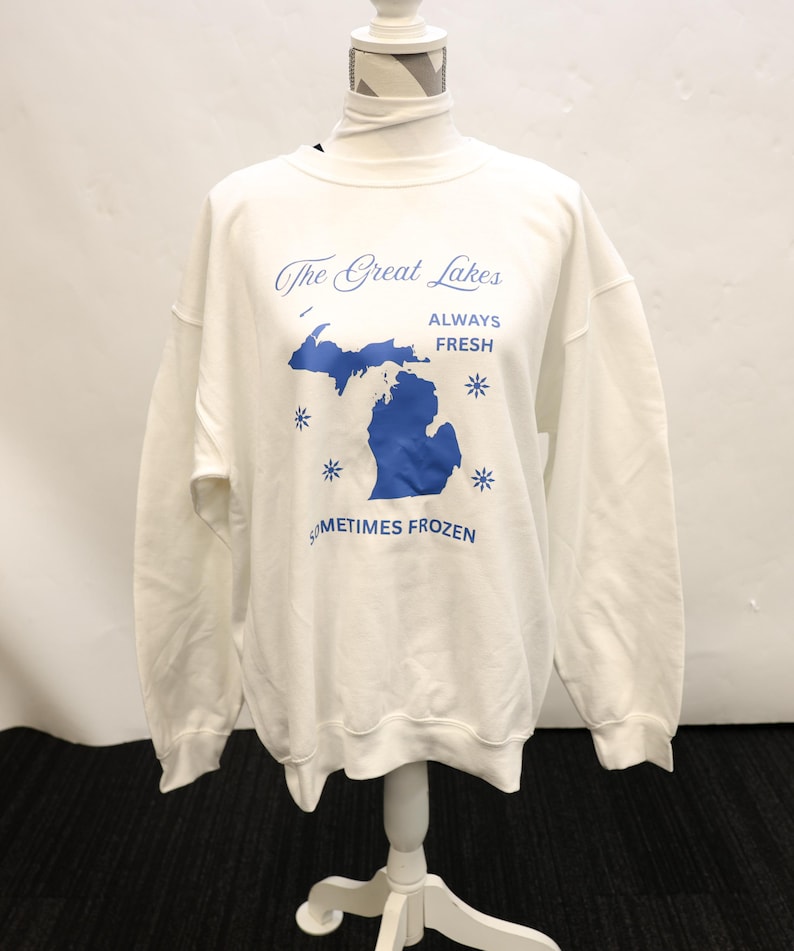 Pu&ograve; includere: Felpa bianca con grafica blu della regione dei Grandi Laghi e la scritta "The Great Lakes ALWAYS FRESH SOMETIMES FROZEN". La felpa &egrave; esposta su un manichino.
