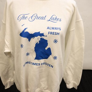 Pu&ograve; includere: Felpa bianca con testo blu e contorno blu dei Grandi Laghi. Il testo recita "The Great Lakes ALWAYS FRESH SOMETIMES FROZEN" con grafica a fiocco di neve. Un top casual e confortevole.
