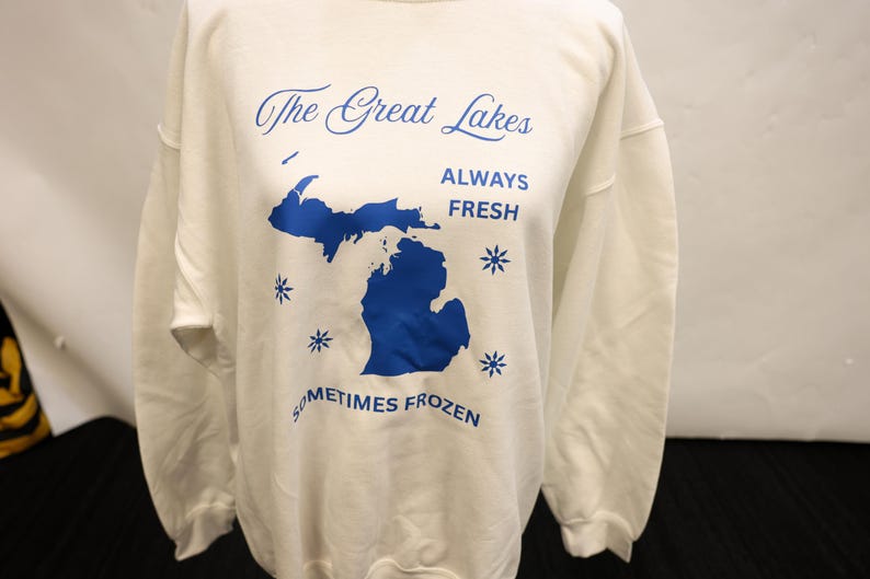 Pu&ograve; includere: Felpa bianca con testo blu e contorno blu dei Grandi Laghi. Il testo recita "The Great Lakes ALWAYS FRESH SOMETIMES FROZEN" con grafica a fiocco di neve. Un top casual e confortevole.
