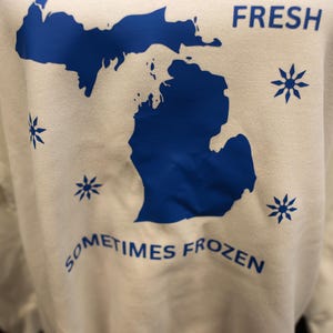 Pu&ograve; includere: Una felpa bianca con un contorno blu dello stato del Michigan. Le parole "FRESH" e "SOMETIMES FROZEN" sono stampate in blu. Disegni di fiocchi di neve blu sono sparsi intorno al contorno dello stato.