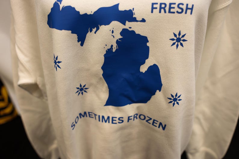 Pu&ograve; includere: Una felpa bianca con un contorno blu dello stato del Michigan. Le parole "FRESH" e "SOMETIMES FROZEN" sono stampate in blu. Disegni di fiocchi di neve blu sono sparsi intorno al contorno dello stato.