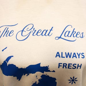 Pu&ograve; includere: Felpa color crema con la scritta "The Great Lakes" in blu sopra una grafica blu della regione dei Grandi Laghi. Sotto la grafica, le parole "ALWAYS FRESH" sono stampate in blu.