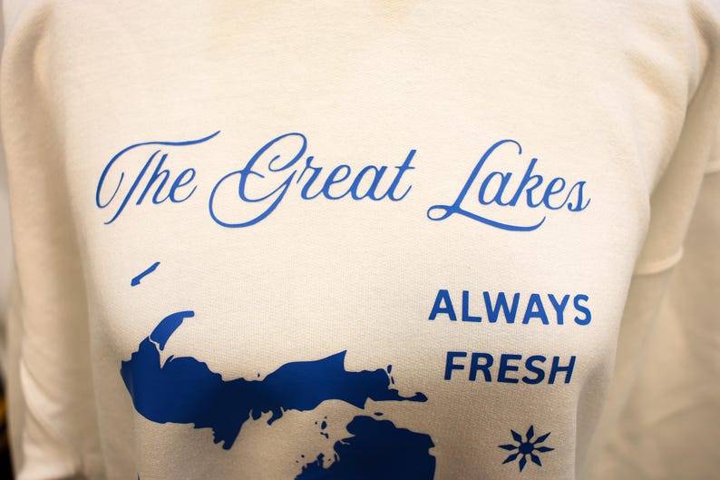 Pu&ograve; includere: Felpa color crema con la scritta "The Great Lakes" in blu sopra una grafica blu della regione dei Grandi Laghi. Sotto la grafica, le parole "ALWAYS FRESH" sono stampate in blu.