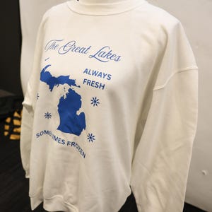 Pu&ograve; includere: Felpa bianca con grafica blu dei Grandi Laghi, incluso lo stato del Michigan. Il testo recita "The Great Lakes" sopra la mappa, con "ALWAYS FRESH" e "SOMETIMES FROZEN" sotto. Sono inclusi anche disegni di fiocchi di neve.