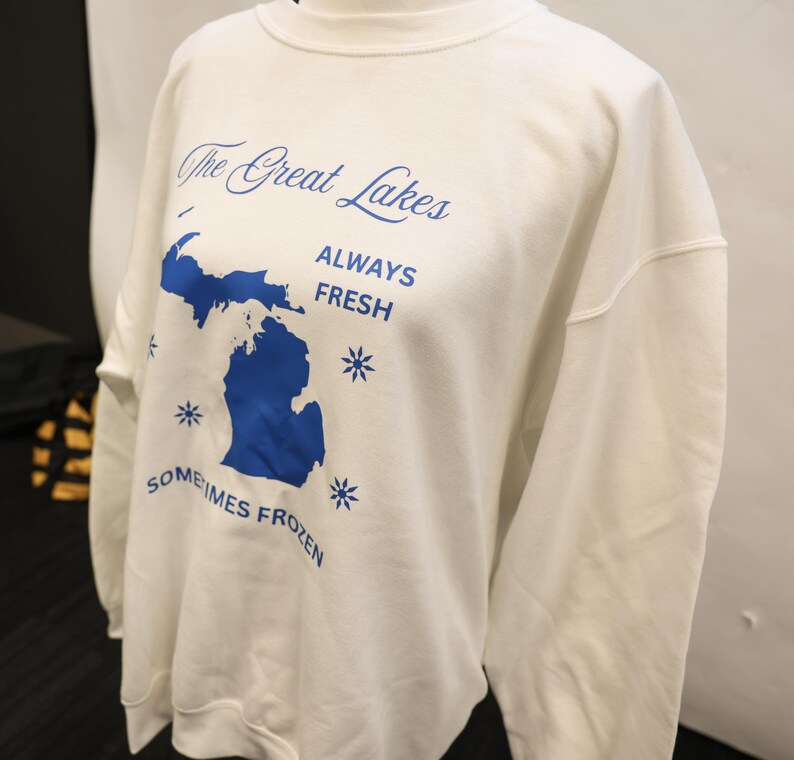 Pu&ograve; includere: Felpa bianca con grafica blu dei Grandi Laghi, incluso lo stato del Michigan. Il testo recita "The Great Lakes" sopra la mappa, con "ALWAYS FRESH" e "SOMETIMES FROZEN" sotto. Sono inclusi anche disegni di fiocchi di neve.
