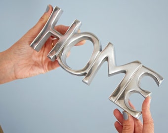 May include: Silver metal home sign with raised letters. The sign is being held by two hands.