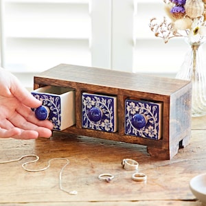 May include: A wooden jewelry box with three drawers. Each drawer has a blue and white floral tile front with a round blue knob. The box is on a wooden surface with jewelry and a lit candle.