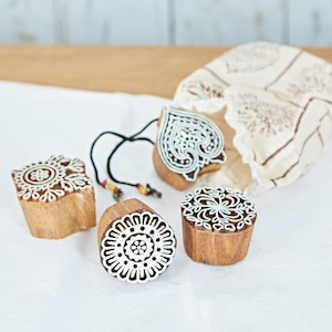 May include: Four wooden block stamps with intricate white designs, including floral and heart shapes. A drawstring bag with a matching pattern sits behind the stamps. The stamps are made of natural wood.