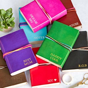 May include: A collection of colourful leather-bound journals with gold lettering. The journals have different titles, including "Dream Big", "Best Mum", "Daddy Cool", "Notes", "My", "Great Ideas", "R.G.B", and "Mouse".