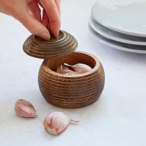 K&ouml;nnte beinhalten: Ein Knoblauchbeh&auml;lter aus Holz mit Deckel, der ein gerilltes Design aufweist. Der Beh&auml;lter ist mit Knoblauchzehen gef&uuml;llt, und daneben liegen drei Zehen auf dem Tisch.